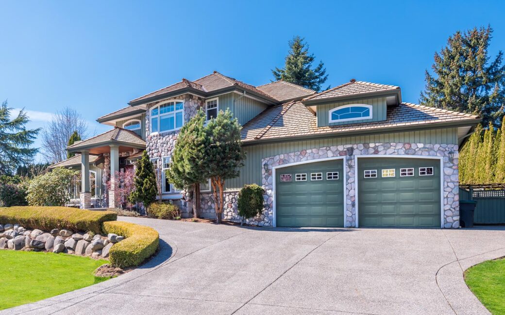 This inviting, eye-level shot presents a suburban house in daylight. The home features a two-car garage and a well-maintained lawn with trimmed bushes and various trees. A curved driveway leads up to the house, under a clear blue sky.