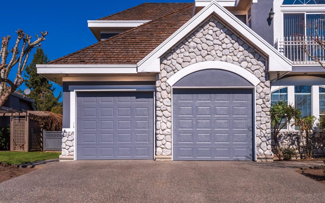 A view of a suburban house on a sunny day. The home features two garage doors, stone accents, and a brown shingled roof.
