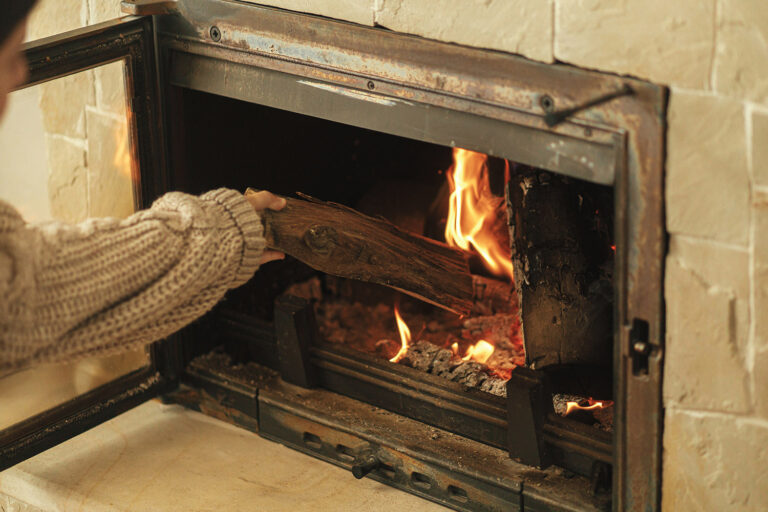 Woman putting firewoods into burning fireplace in rustic room in farmhouse. Heating house in winter with wood burning stove. Fireplace heating alternative to gas and electricity